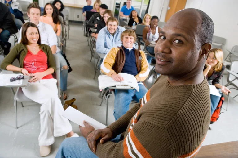 A male teacher faces the camera and smiles while standing in front of a classroom